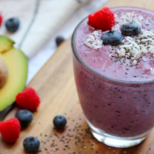purple fertility smoothie in a glass with decorative avocado and berries in background