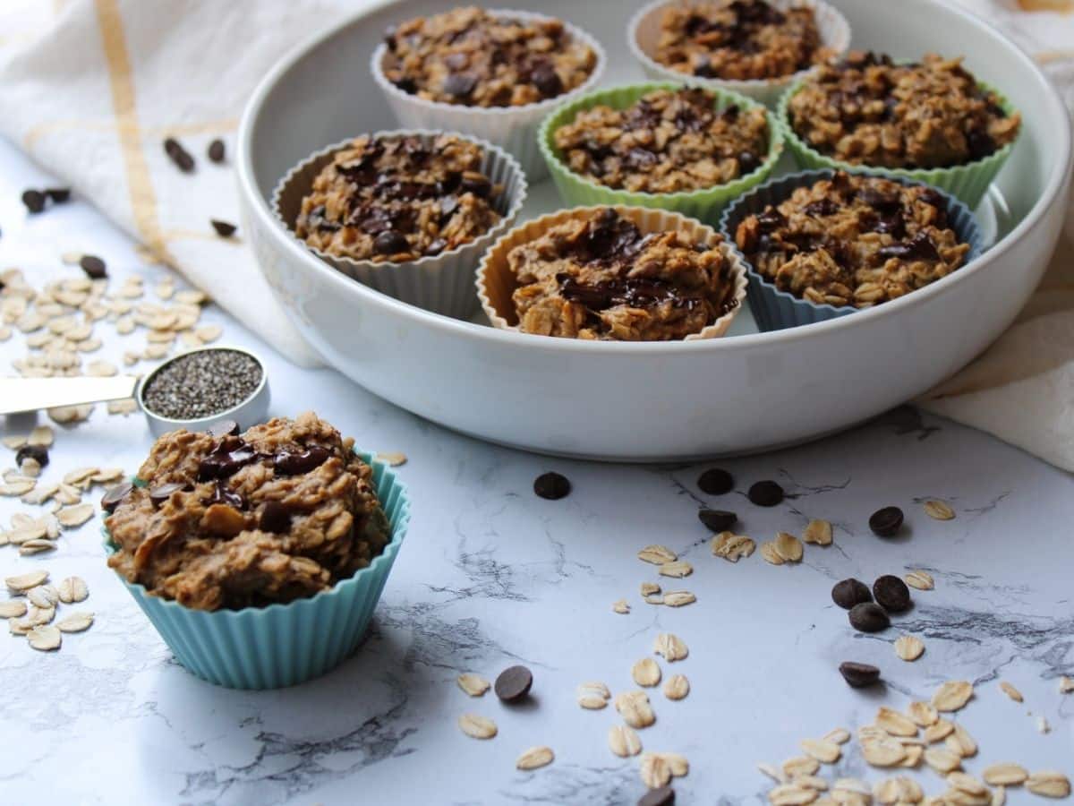 Photo of a peanut butter banana oatmeal cup on a white countertop with a plate of additional muffins in the background. Oats and chocolate chips are spread along the countertop. 