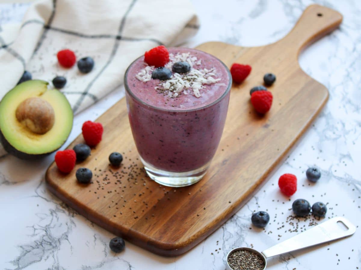 Purple fertility smoothie on a cutting board with avocado, berries, and chia seeds decoratively placed in background on countertop