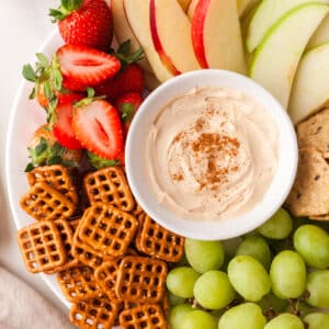 pb yogurt dip in white bowl on a platter with fruit and pretzels surrounding it.
