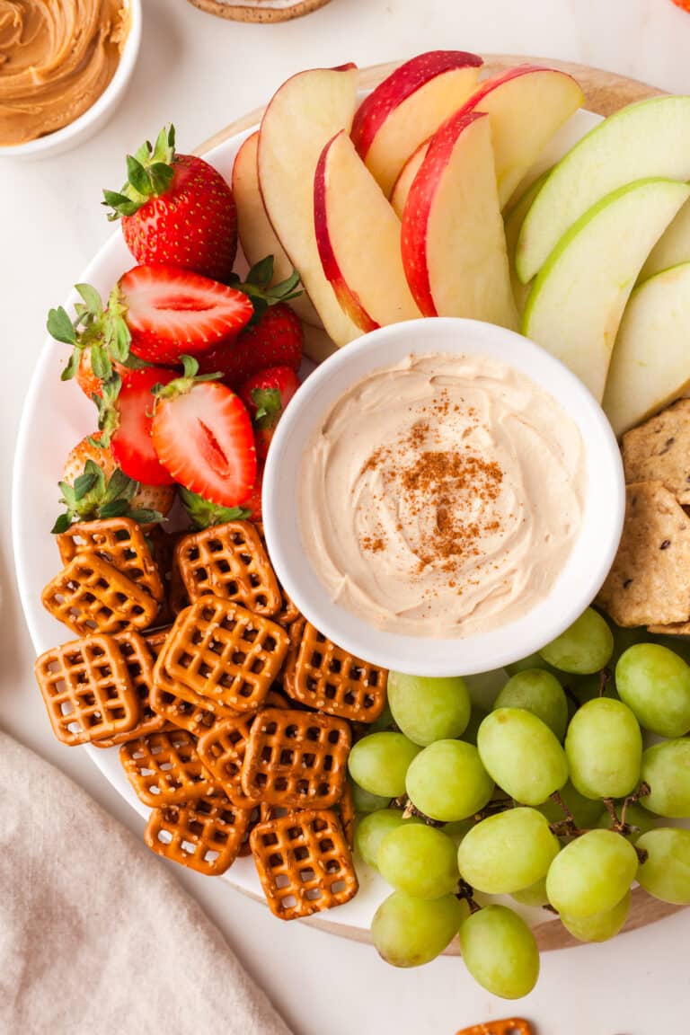 pb yogurt dip in white bowl on a platter with fruit and pretzels surrounding it.