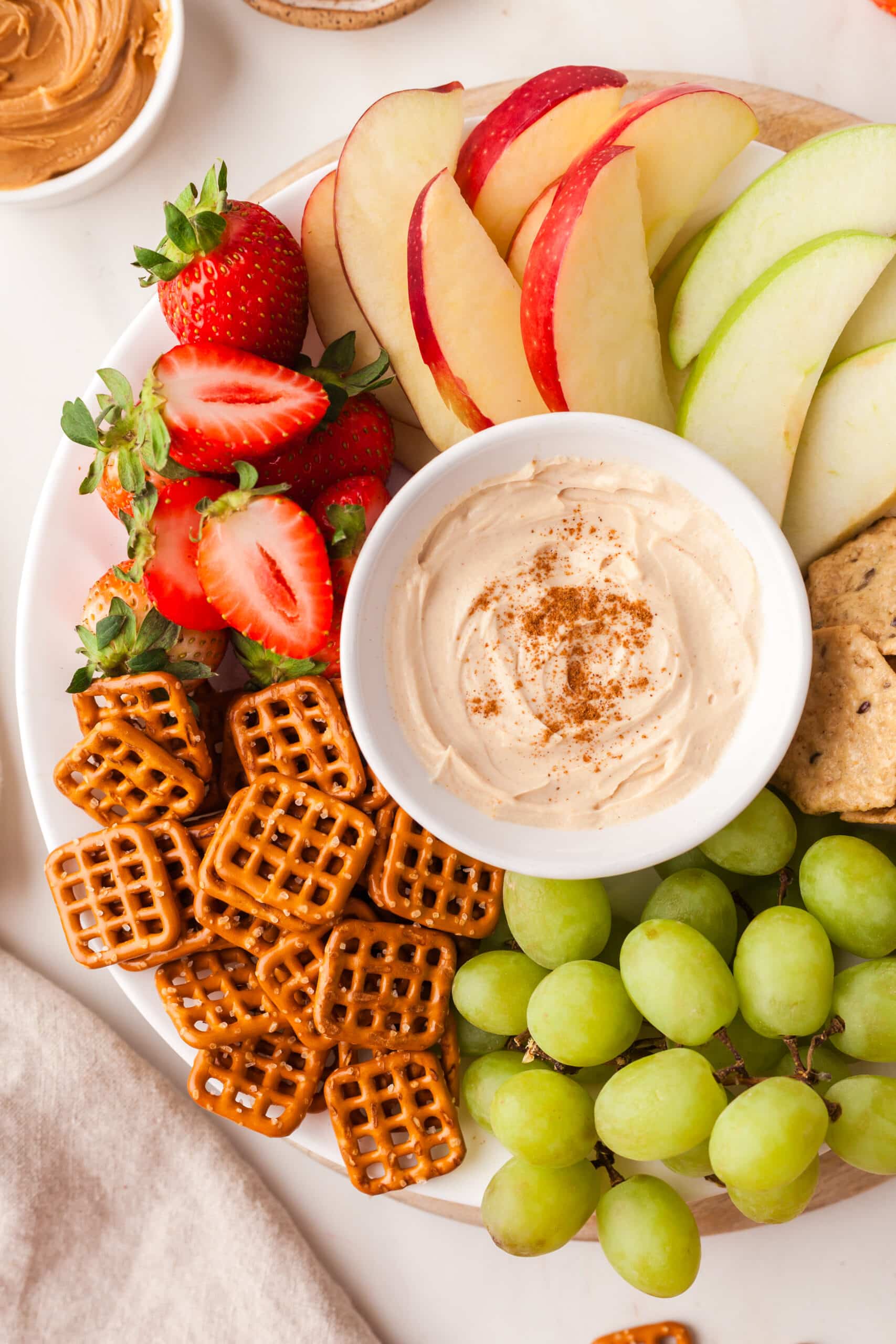 pb yogurt dip in white bowl on a platter with fruit and pretzels surrounding it.