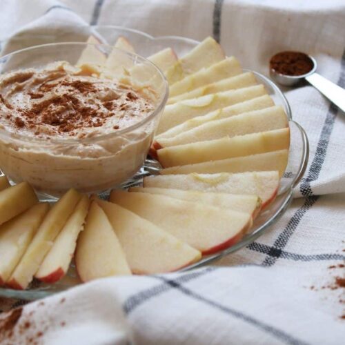 Photo of peanut butter yogurt dip in a bowl sprinkled with cinnamon on top. Sliced apples surround the dip bowl.