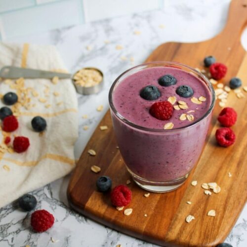 triple berry oat smoothie in a glass on a cutting board with oats and berries spread around it.