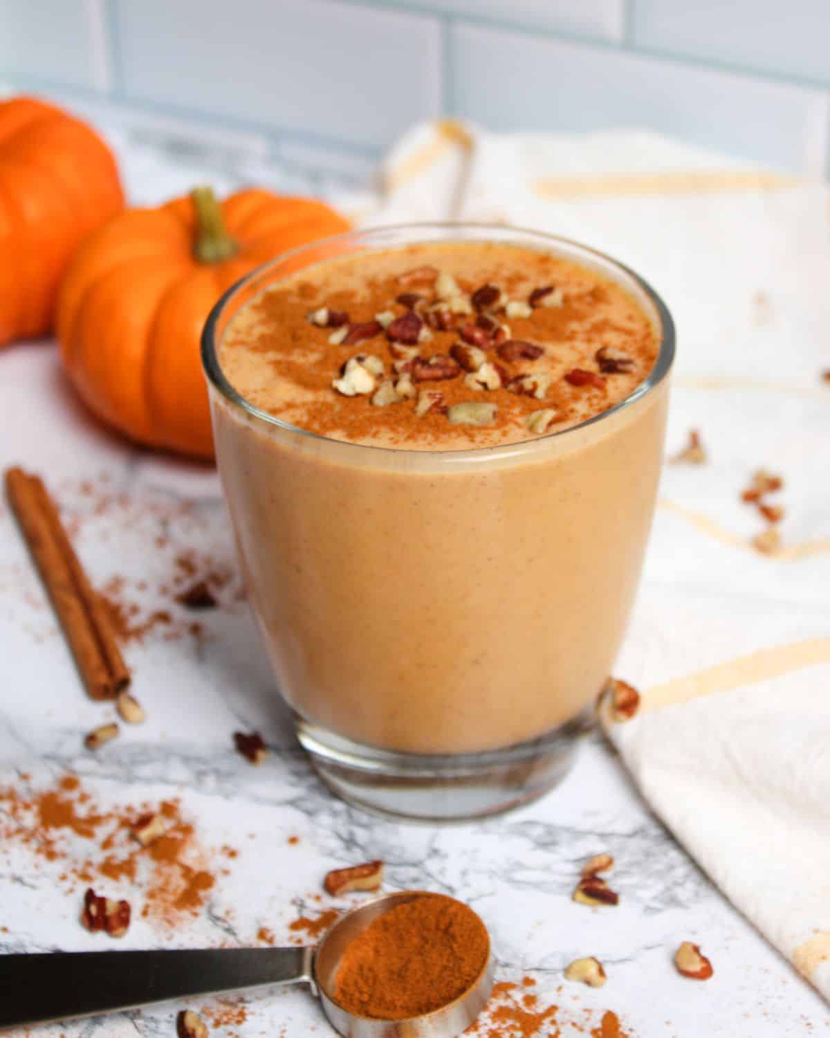 pumpkin smoothie in a glass with cinnamon and chopped nuts on top. pumpkins and cinnamon sticks in the background