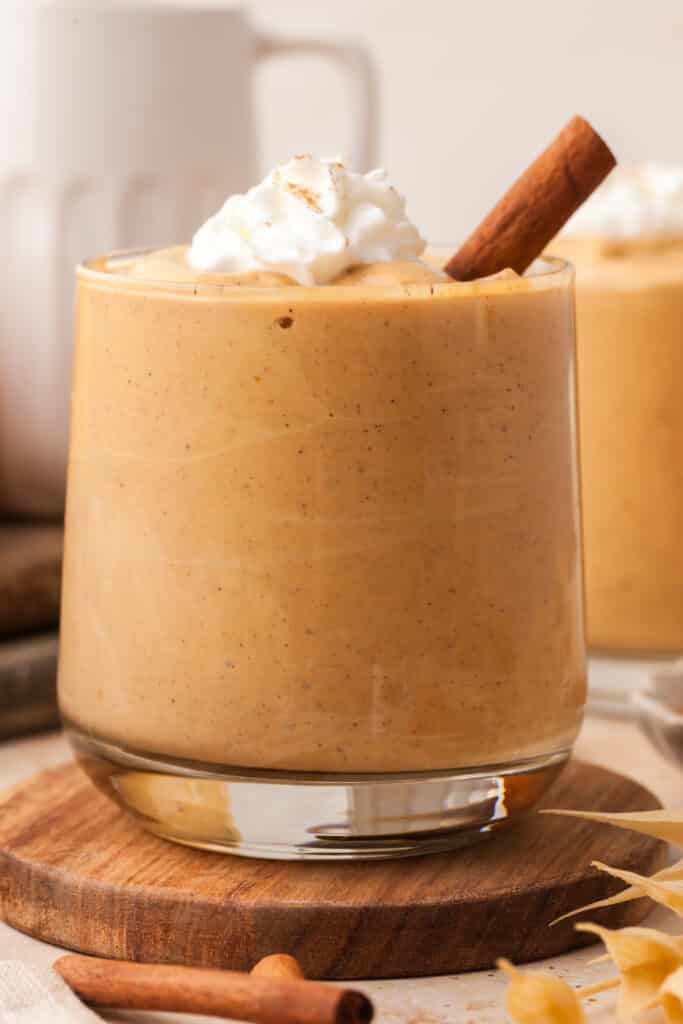 protein pumpkin smoothie in a glass with whipped topping and a cinnamon stick.