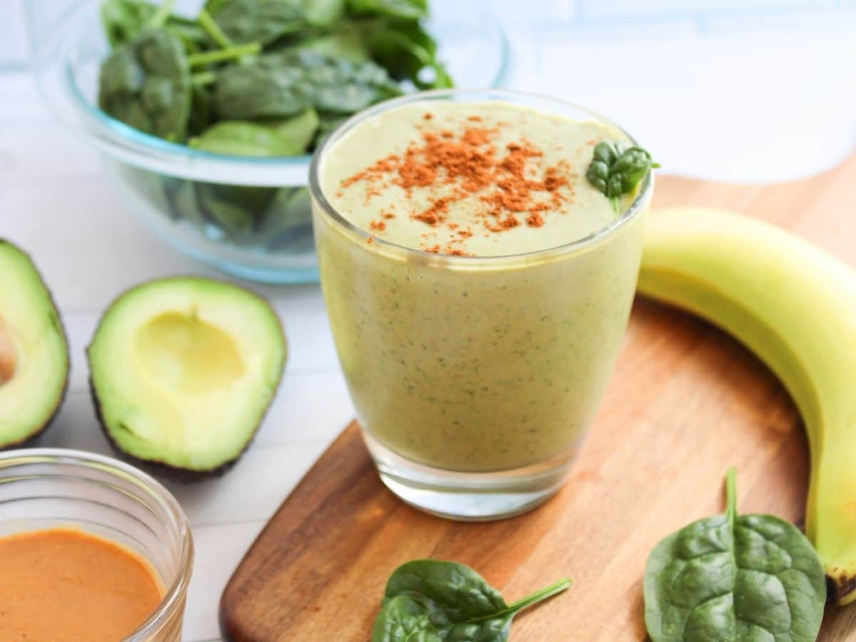 peanut butter avocado smoothie in a glass with a bowl of spinach, banana, open avocado, and bowl of peanut butter decoratively surrounding the smoothie glass