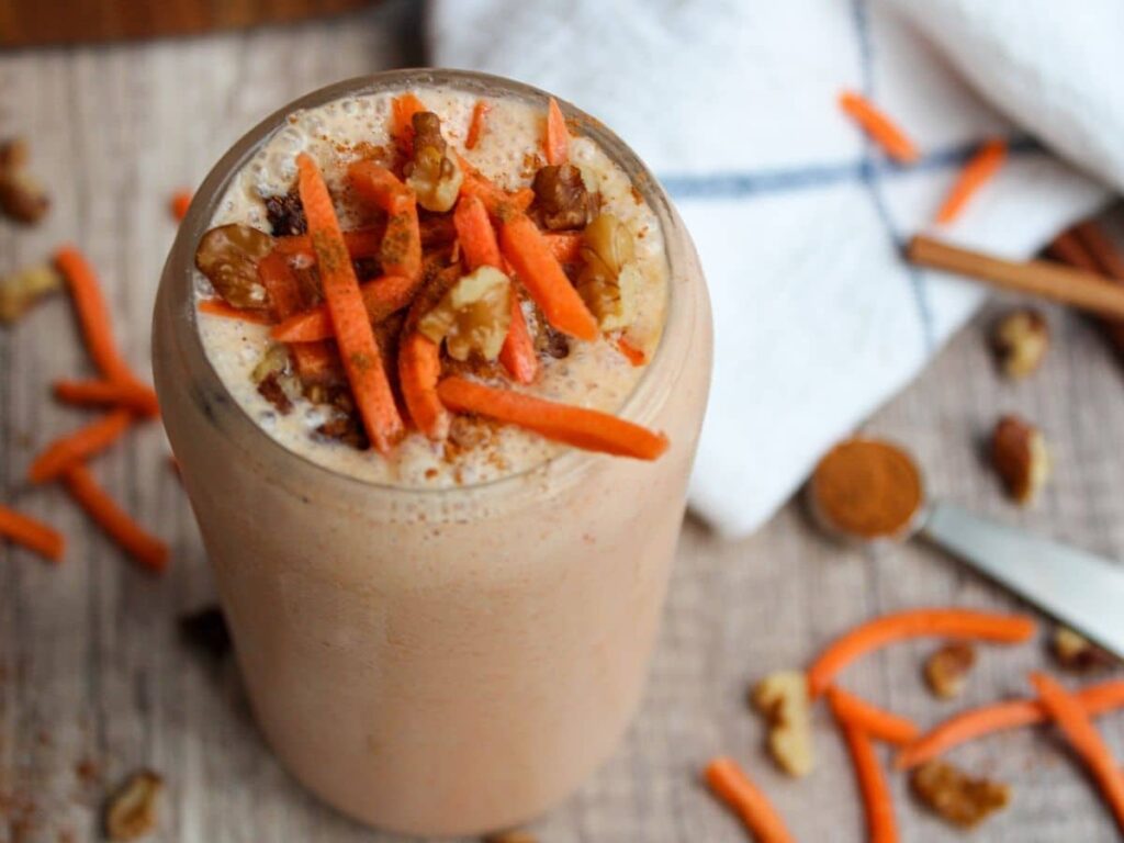 carrot banana smoothie in a glass on a wooden surface. Decorative carrots and cinnamon in the background.