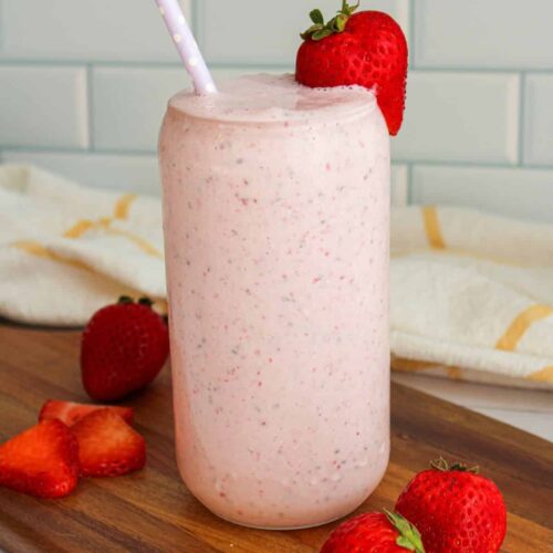 vertical view of a pink strawberry smoothie in a tall glass with a strawberry perched on the rim and a purple straw coming out. The smoothie glass is on a cutting board with strawberries laid out.