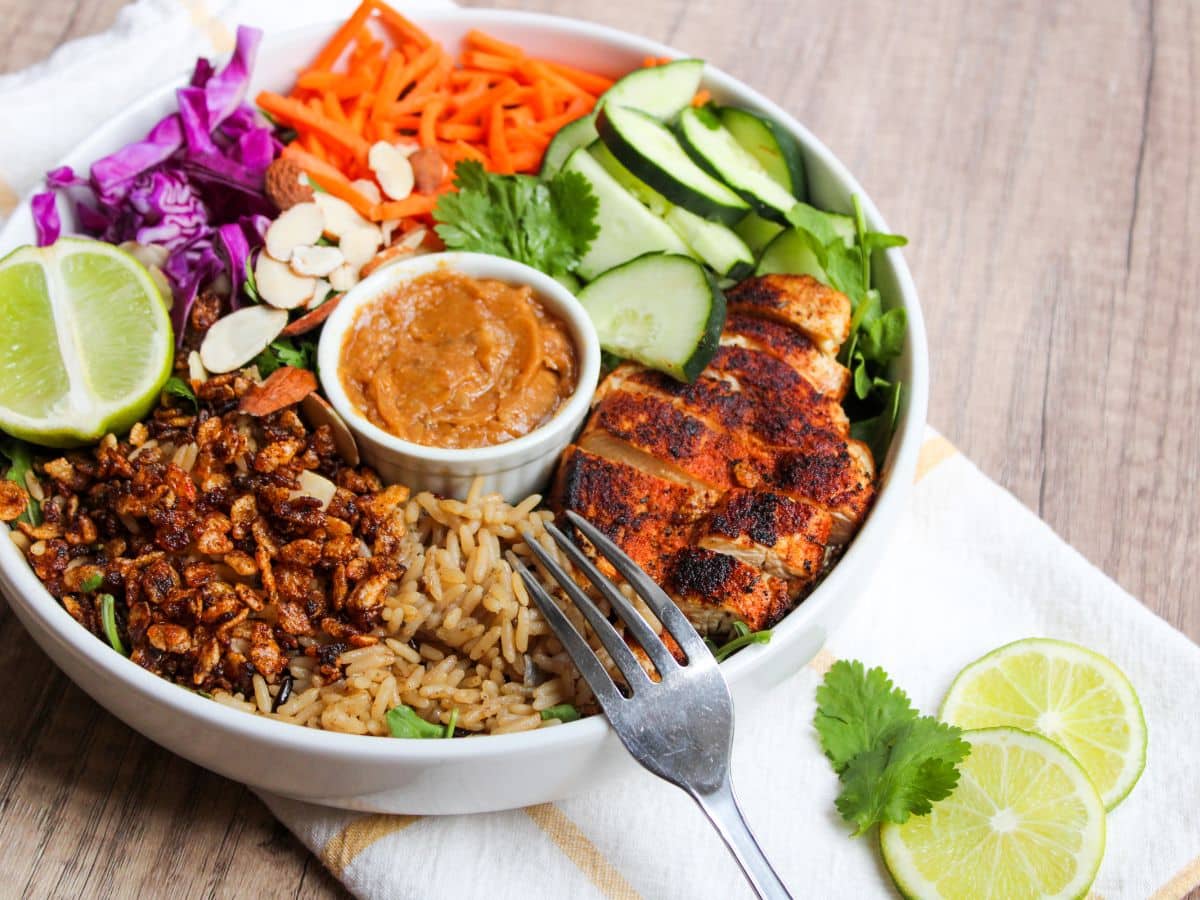 blackened chicken, wild rice, crispy rice, veggies, and spicy cashew dressing on a plate. Plate is sitting on top of a napkin with a fork and decorative lime slices.