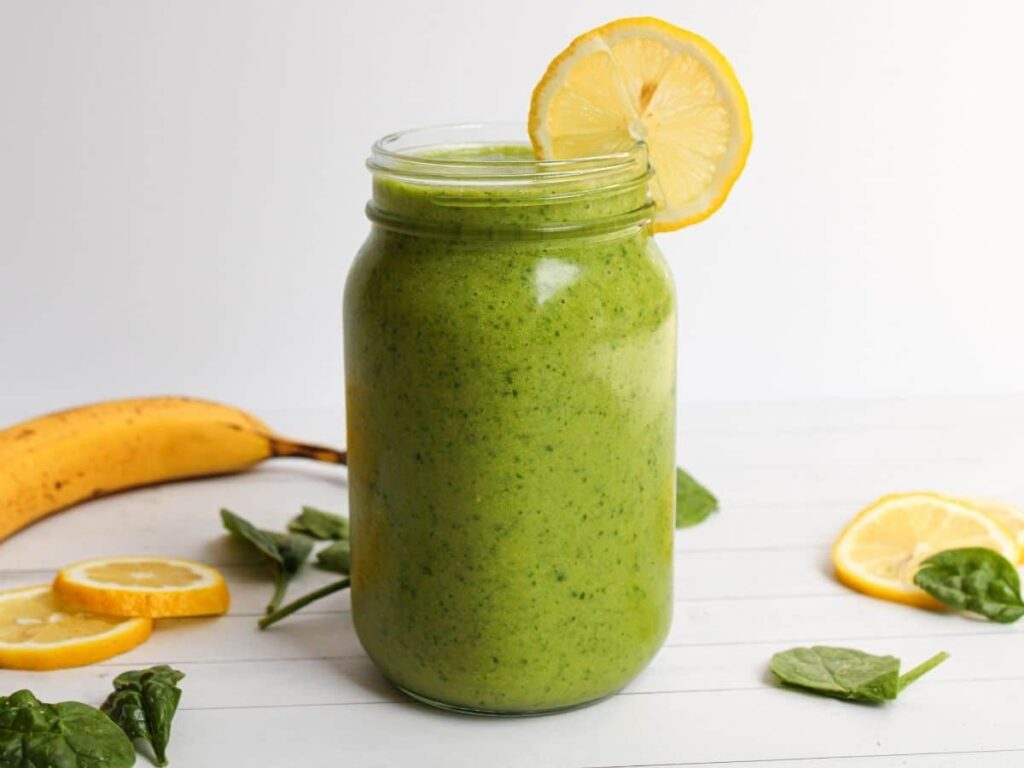 Detox Island Green Smoothie in a mason jar with a banana, spinach leaves, and lemon wedges decorative in the background.