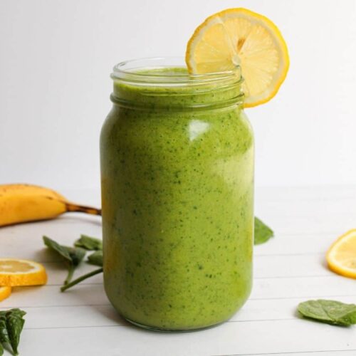 Detox Island Green Smoothie in a mason jar with a banana, spinach leaves, and lemon wedges decorative in the background.
