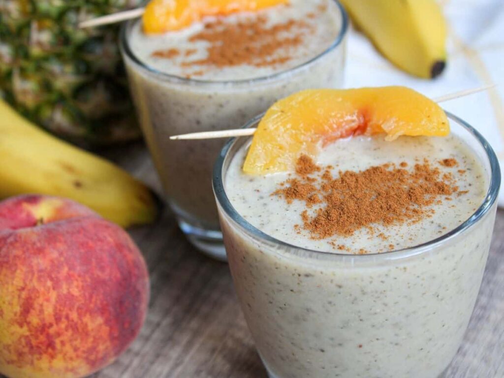 peach pineapple smoothie in a glass with fruit in the background and a peach slice and cinnamon on top of the smoothie.