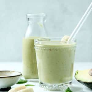 peanut butter avocado smoothie in a glass with glass straw and ingredients surrounding it.
