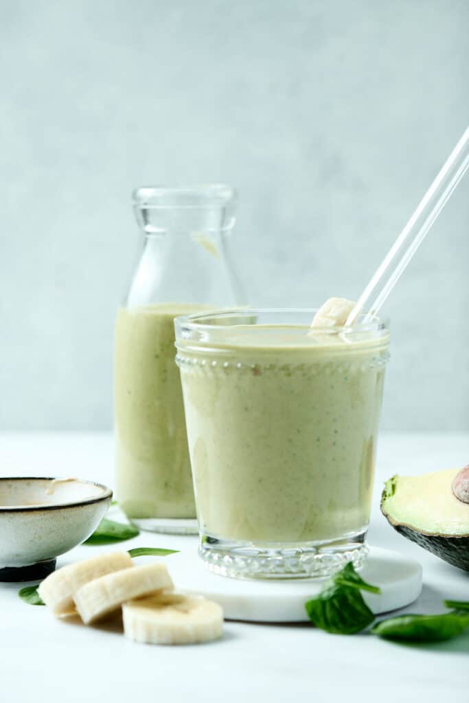 peanut butter avocado smoothie in a glass with glass straw and ingredients surrounding it.