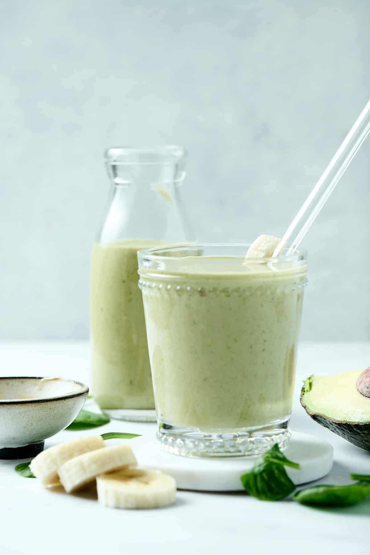 peanut butter avocado smoothie in a glass with glass straw and ingredients surrounding it.