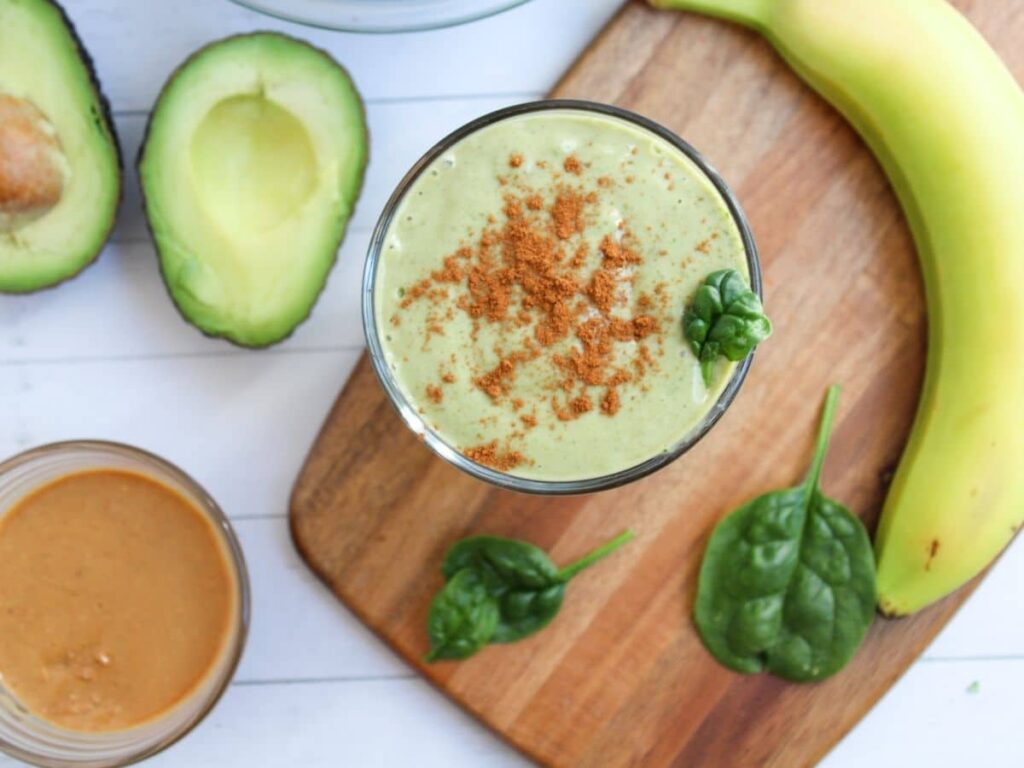 overview photo of a green avocado smoothie with cinnamon spinkled on top. A banana, open avocado, and bowl of peanut butter are in the background decoratively.