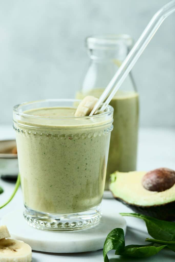 peanut butter avocado smoothie in a glass with glass straw and ingredients surrounding it.