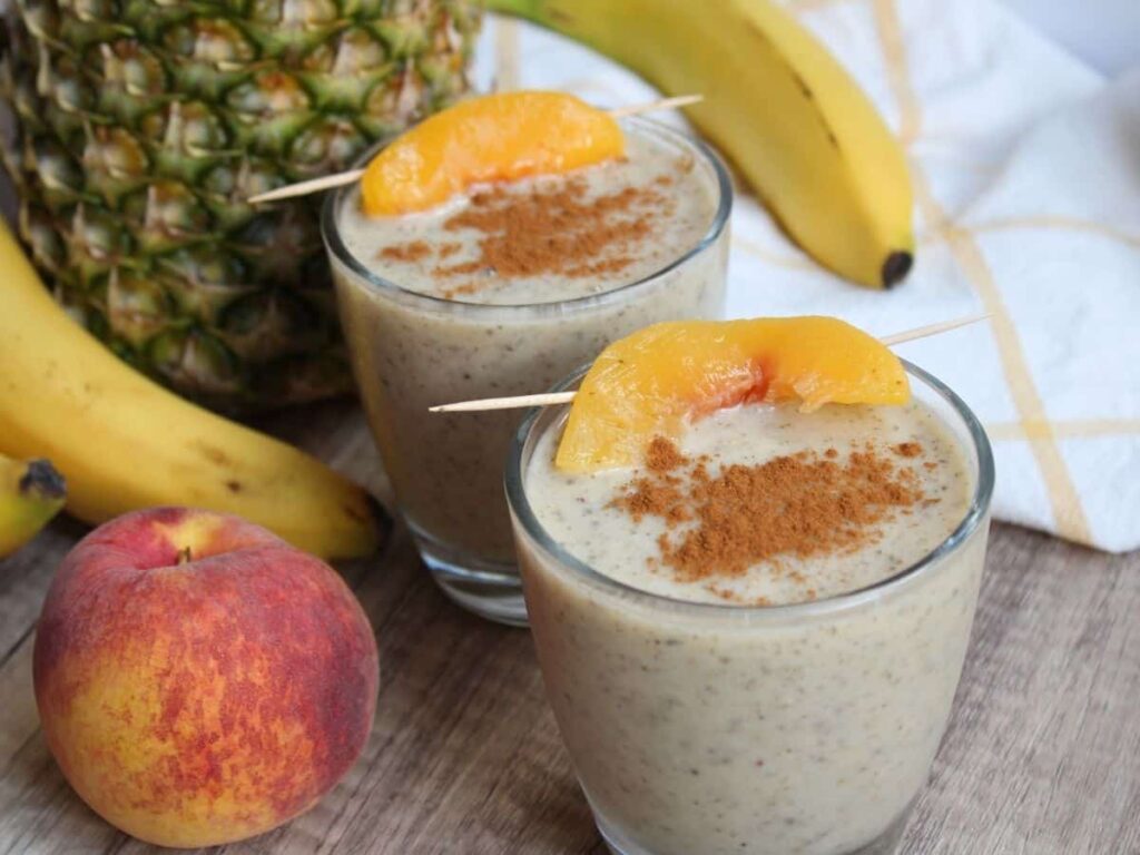 two pineapple peach smoothies in glasses with a peach slice and cinnamon on top. Fruit is in the background.
