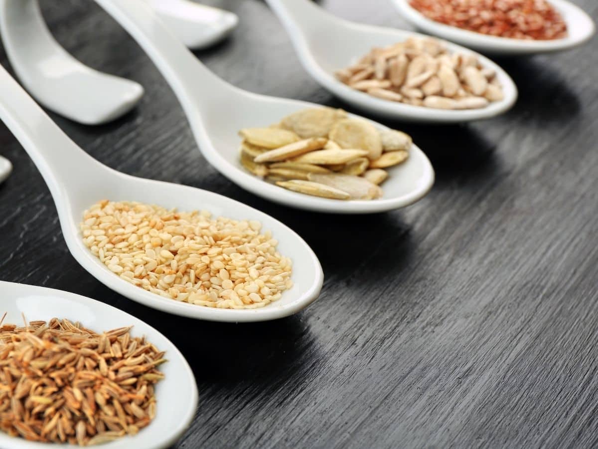 Assorted seeds on white spoons sitting on a wooden table.