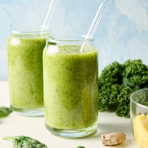 two green detox island tropical smoothies in glasses with straws; kale and other ingredients are on countertop too.