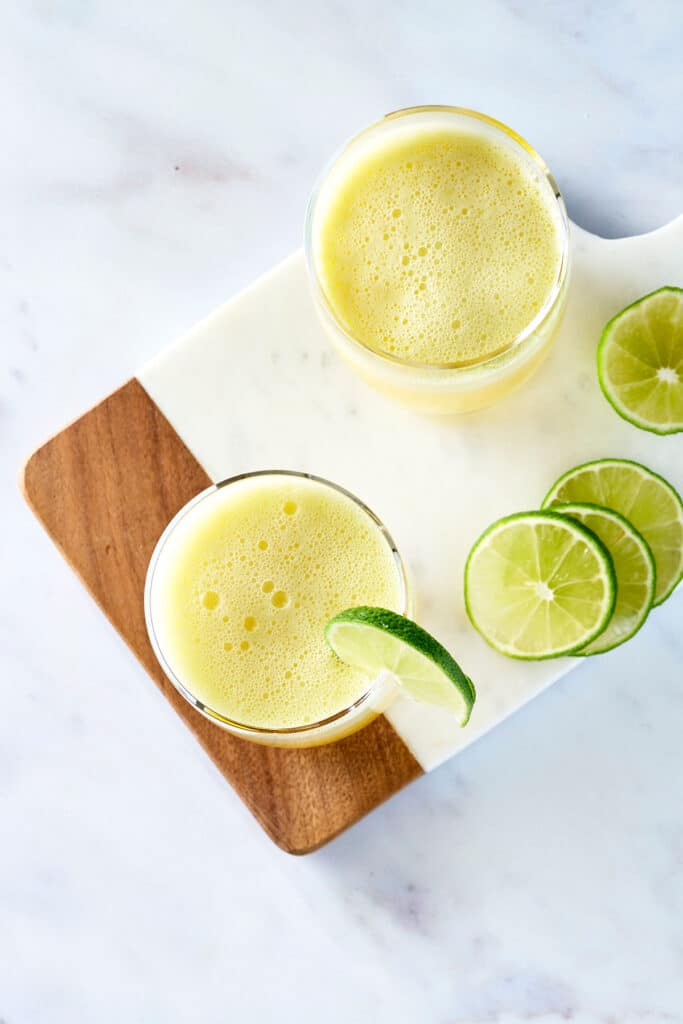 overview of two pineapple mocktails in glasses with limes on a wood cutting board.