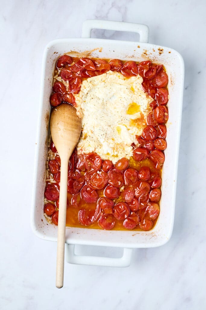 roasted cherry tomatoes and feta in a baking dish with wooden spoon.