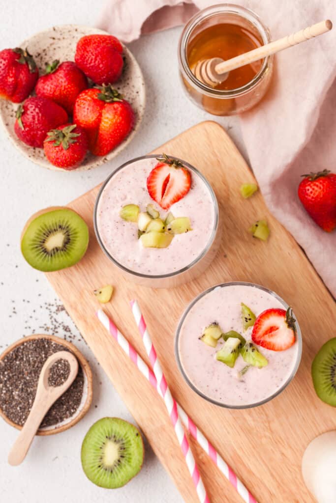 pink colored smoothies in two glasses with strawberry and kiwi garnish and a straws beside them.