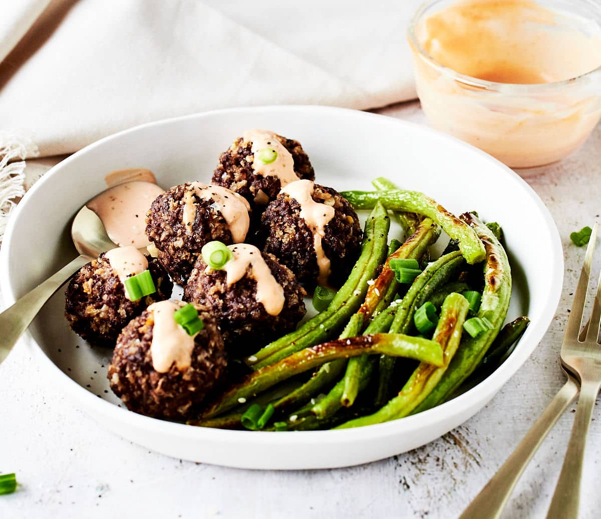 firecracker meatballs with a sauce drizzled on top with green beans in a white bowl.