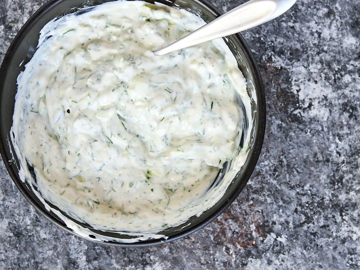 homemade tzatziki sauce in a bowl on dark countertop with a spoon in it.