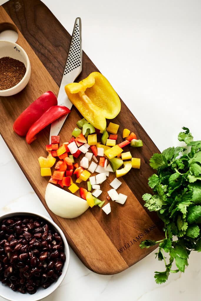 overhead shot of chopped bell peppers, onions, taco seasoning, and cilantro on a wooden cutting board next to a bowl of black beans.
