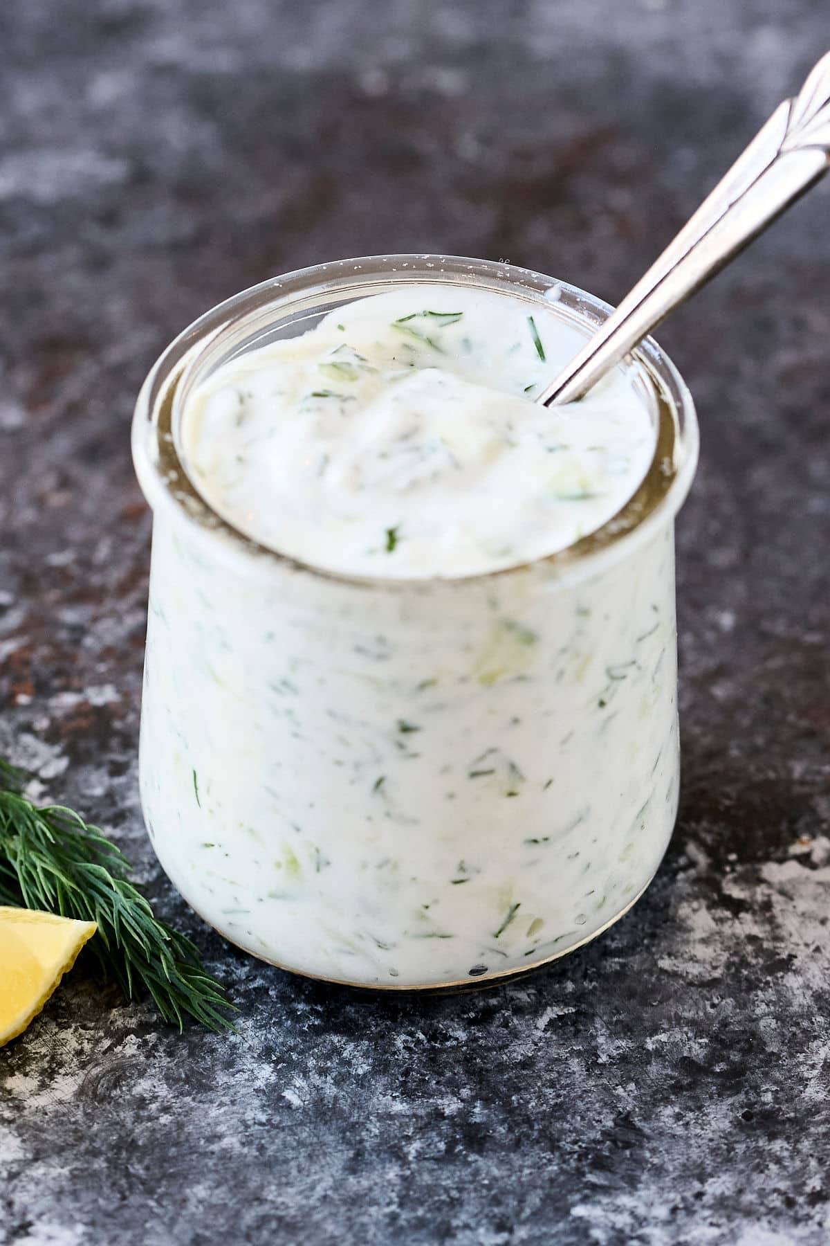 homemade tzatziki sauce in a glass jar on dark countertop with a spoon in it.