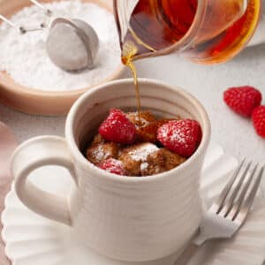 french toast in a white mug recipe with powdered sugar and raspberries on top; maple syrup is being poured onto it.