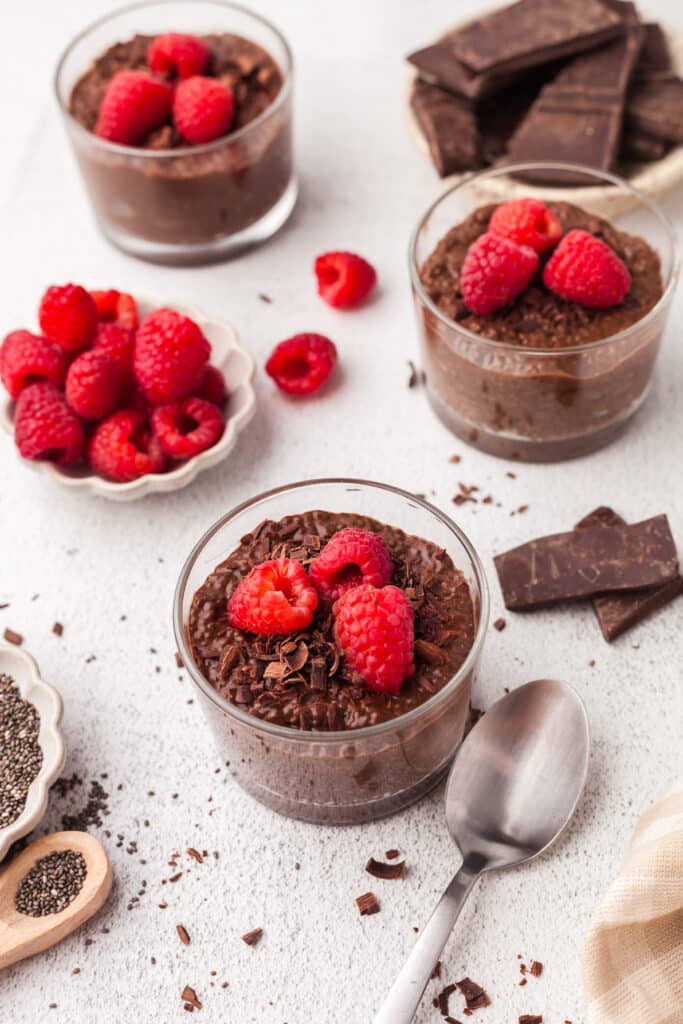 chocolate chia puddings in glass jars on a countertop with chocolate and raspberries decoratively placed.