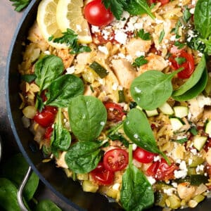 chicken orzo bake in a skillet with fresh herbs and lemon wedges.