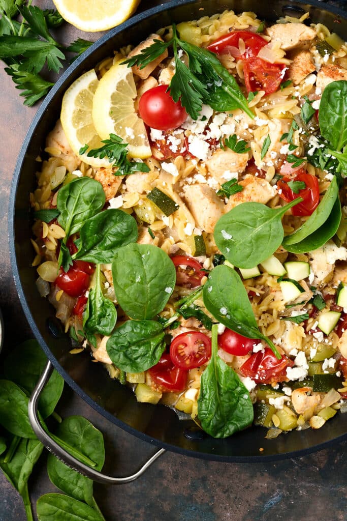 chicken orzo bake in a skillet with fresh herbs and lemon wedges.