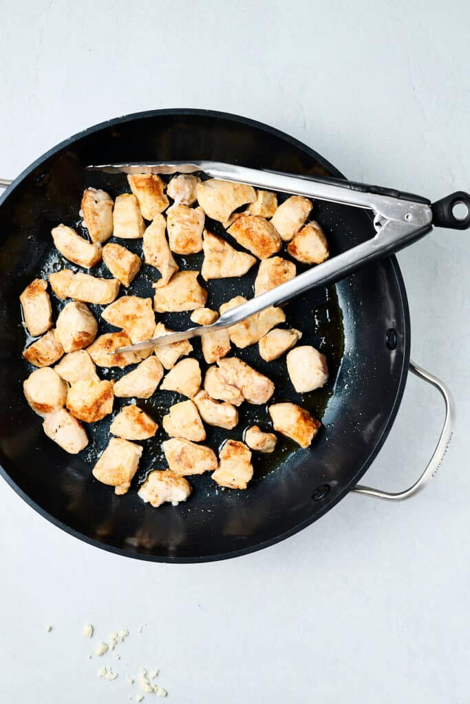 chicken cubes sautéing in a skillet with tongs.