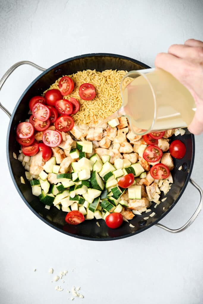 pouring chicken broth into a chicken orzo bake skillet.