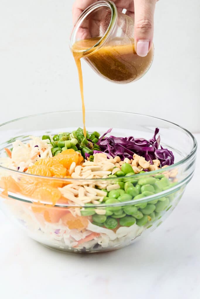 ingredients for a crunch asian salad in a large glass bowl with sesame dressing being poured over.