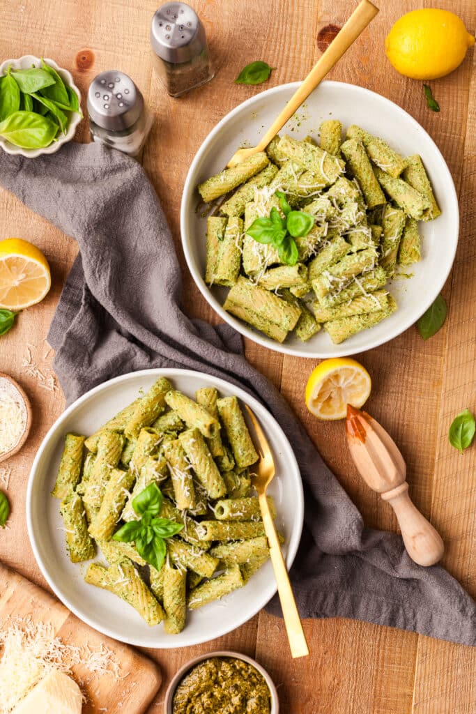 protein pesto pasta in white bowls with gold spoons inserted on wooden countertop.
