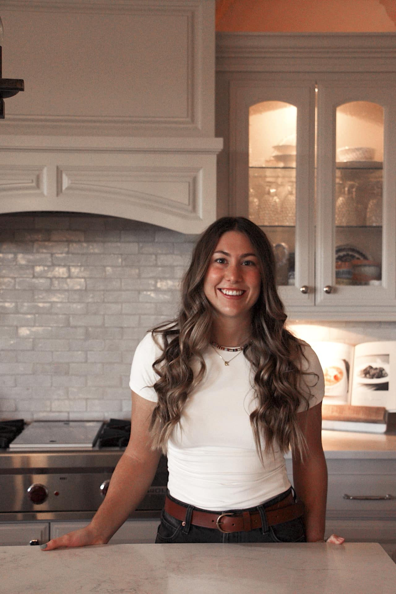 woman with a white top and long brunette hair smiling at camera in the kitchen.