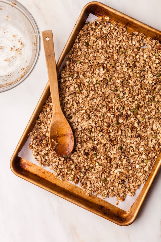 granola mixture being spread onto parchment lined baking sheet with wooden spoon.