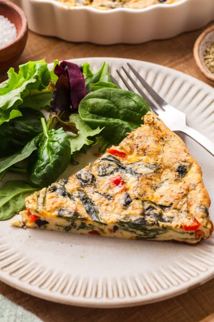 slice of crustless quiche on a white plate with greens beside it.