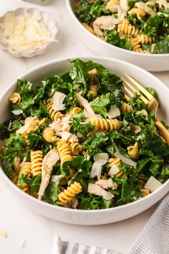kale pasta caesar salad in a white bowl with gold fork inserted.