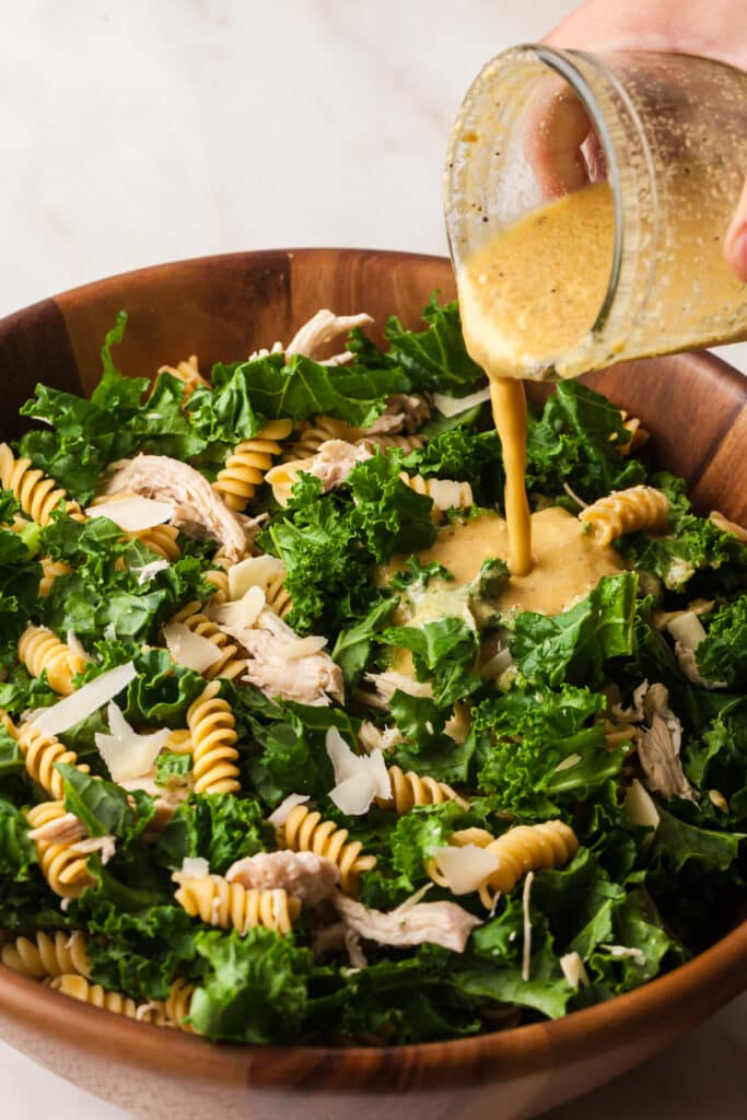 Salad dressing being poured from glass jar onto kale pasta caesar salad in wooden bowl.