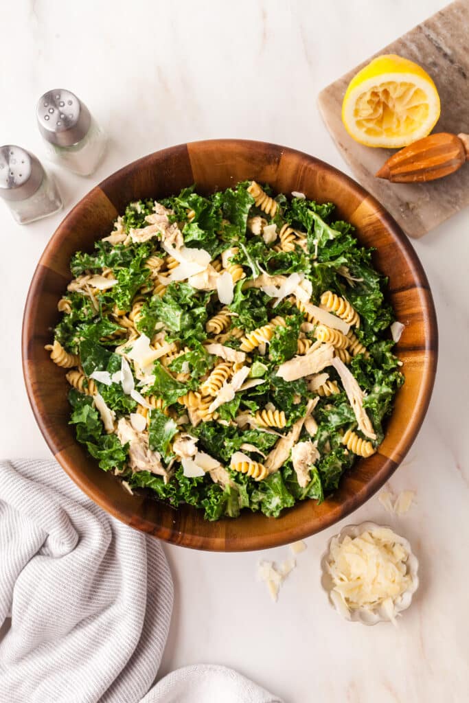 kale pasta caesar salad in a wooden bowl with ingredients surrounding it.